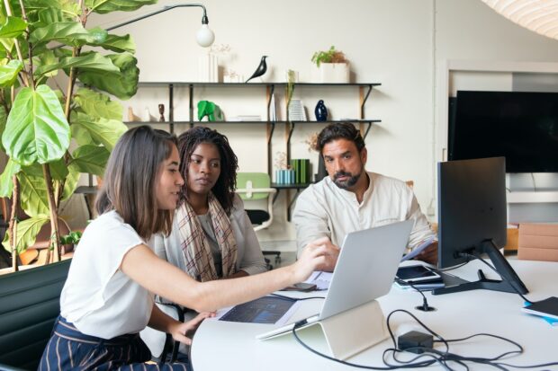 A group of three office workers, two women and one man, are looking at a laptop. One woman is describing something on the laptop.