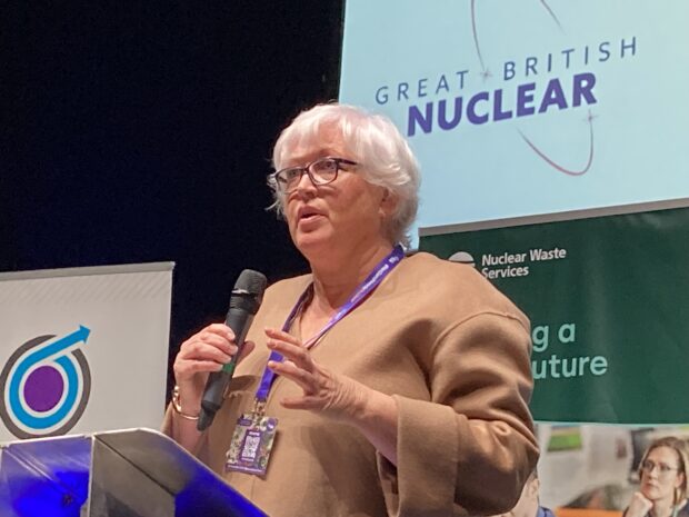 Gwen Parry-Jones addresses a room of people at the Nuclear Institute YGN Annual Conference. She is holding a mic, addressing people from a stage. A Great British Nuclear logo is behind her, above a Nuclear Waste Services banner
