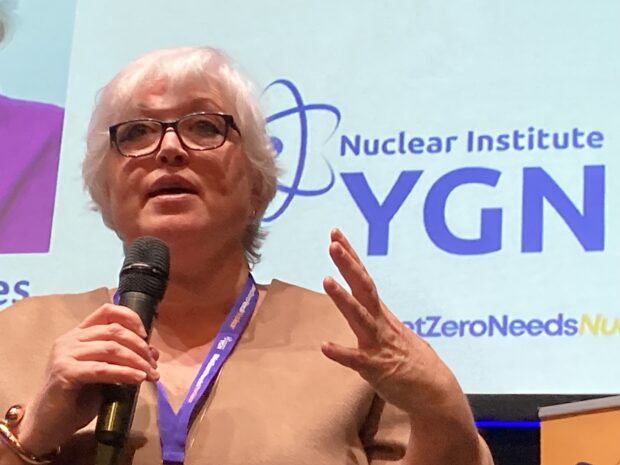Gwen Parry-Jones addresses a room of people at the Nuclear Institute YGN Annual Conference. She is holding a mic, addressing people from a stage. Behind her, the Nuclear Institute YGN sign is visible.