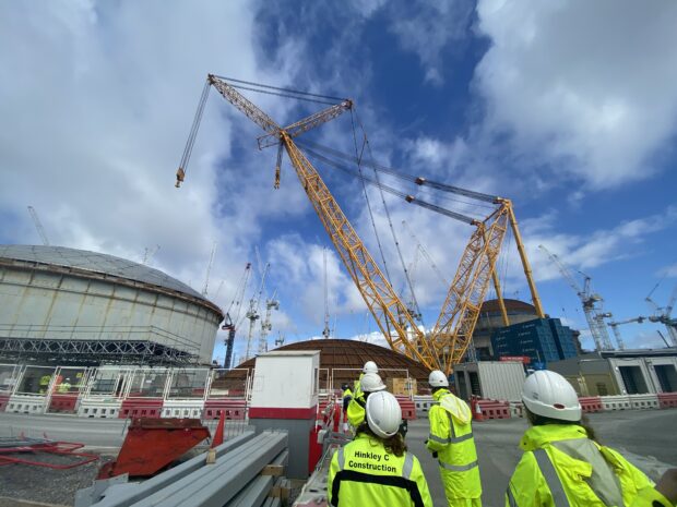 The photo is of Hinkley Point C, a nuclear power station under construction. The photo includes numerous construction cranes. In the foreground of the photo are a group of visitors wearing high-vis yellow jackets and trousers, white hard hats and black boots. In the background of the photo is the site, bordered by red and white traffic safety barriers and white fencing.