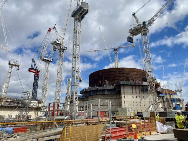 The photo is of Hinkley Point C, a nuclear power station under construction. The photo includes numerous construction cranes. Several large concrete towers are being built. Members of staff in high-vis clothing and white hard hats are working and handling building materials. Above the construction site are a blue sky and white clouds.