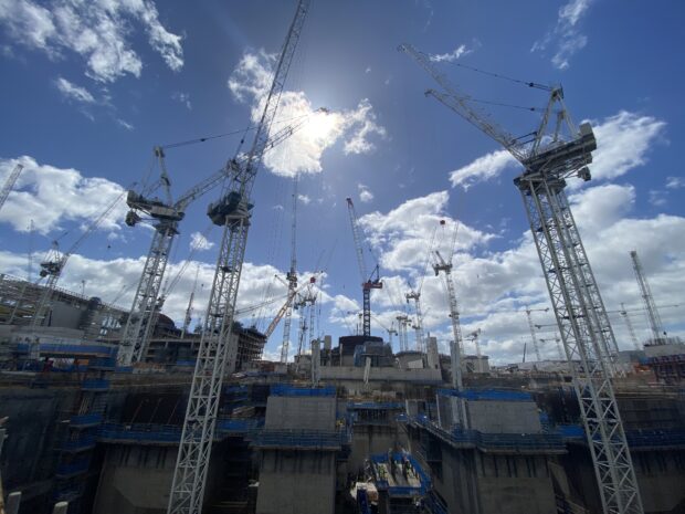 The photo is of Hinkley Point C, a nuclear power station under construction. The photo includes numerous construction cranes. Several large concrete towers are being built. Above the construction site are the sun, a blue sky and white clouds.