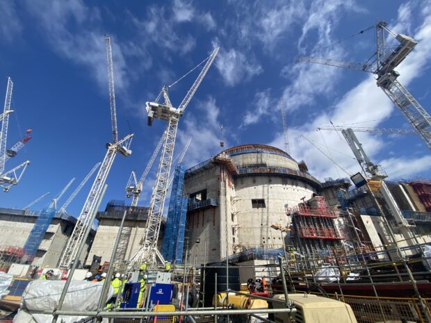 The photo is of Hinkley Point C, a nuclear power station under construction. The photo includes numerous construction cranes. A large dome is being constructed. In the foreground, there are numerous building and construction materials. Staff wearing yellow high vis clothing and white hard hats are working and handling building materials. Above the construction site are a blue sky and white clouds.